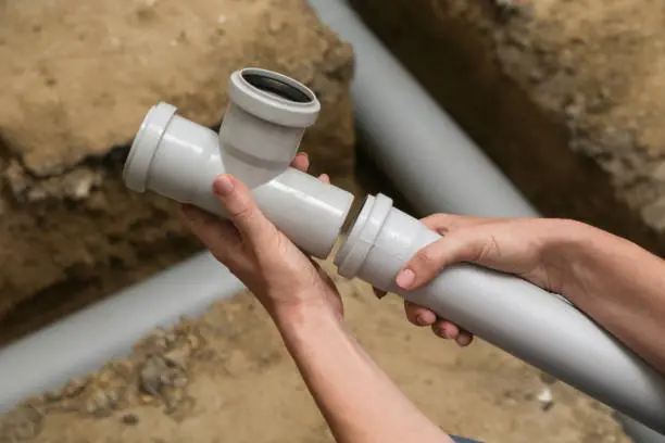 > Hands holding and fitting together grey plastic pipe joints in an open trench, demonstrating how shifting soil affects PVC sewer lines by bending, pulling apart, or fracturing underground plumbing connections.