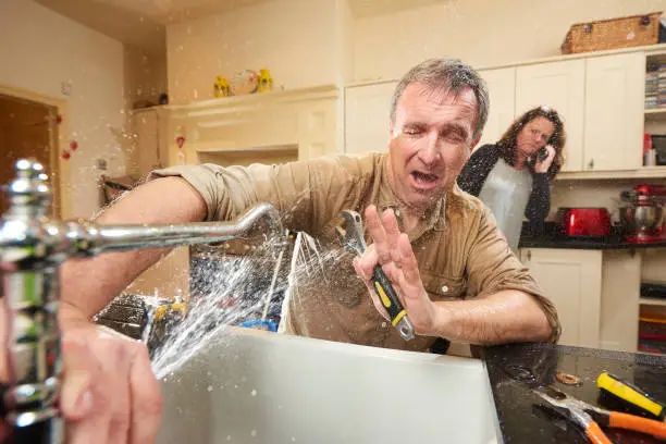 A stressed man getting sprayed with water while trying to learn how to fix a leaky faucet in a kitchen.