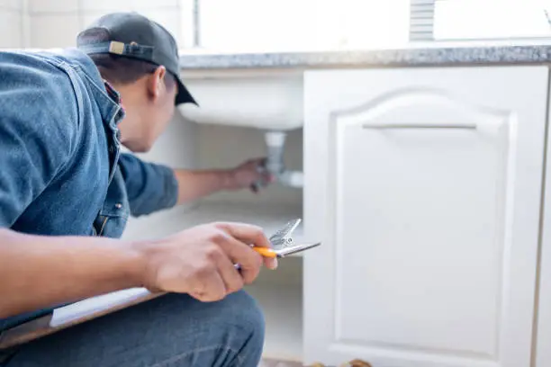 An Oklahoma City plumber checking a residential plumbing inspection checklist while reviewing the drainage pipes under a kitchen sink.