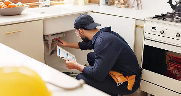 A professional plumber using a plumbing inspection checklist for OKC homes to examine pipes under a kitchen sink.