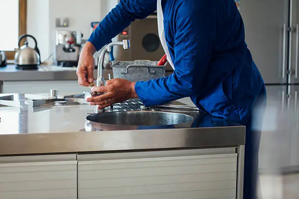 Close-up of a person in blue workwear using hands and tools to repair a kitchen sink fixture, detailing how to fix a leaky faucet.