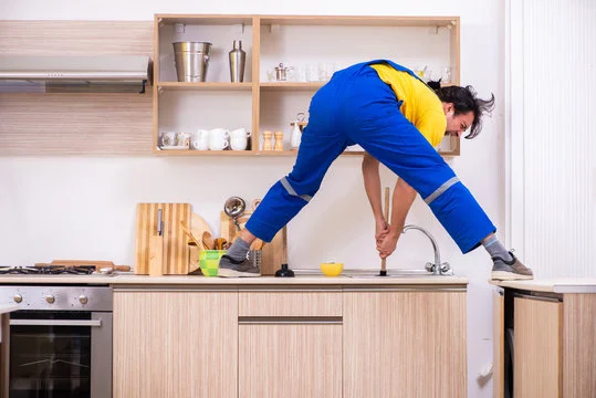 A plumber in blue overalls stretching across a kitchen counter to use a plunger on a sink, demonstrating why faucets start to drip or clog.