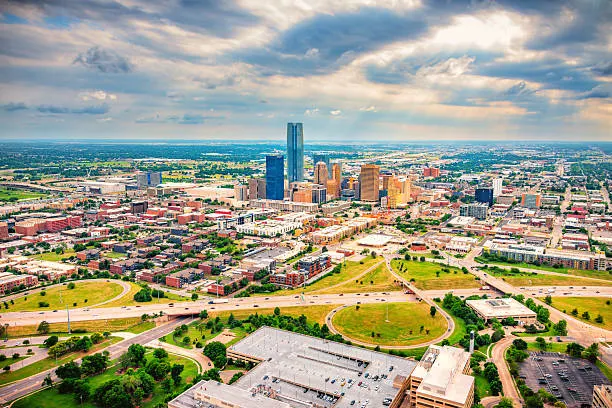 High-angle aerial photograph of the Oklahoma City skyline under a dramatic, cloudy sky, with green landscapes in the foreground. The image provides environmental context for the rapid weather changes that affect OKC homes.