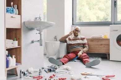 Stressed man sitting on a wet bathroom floor making a call near broken plumbing pipes.