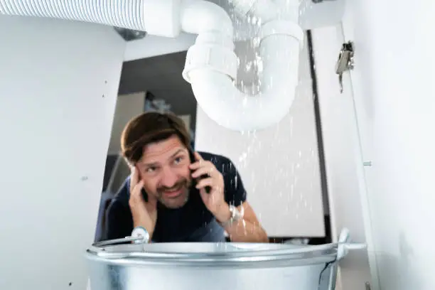 Worried homeowner on phone call looking at leaking PVC pipe under sink during home plumbing restoration.